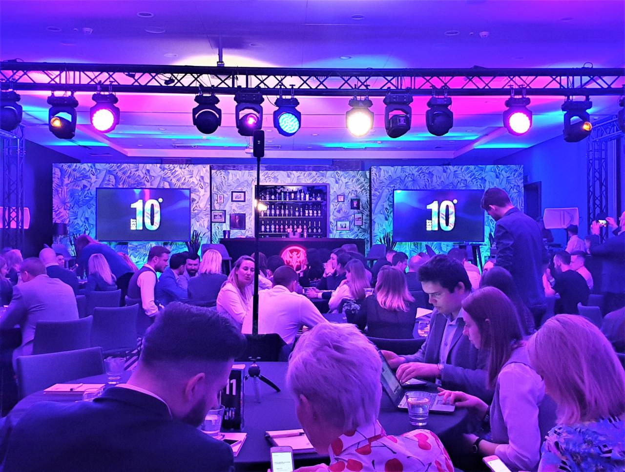 Konferencja w sali z oświetleniem scenicznym, ekranami z numerem 10, stołami i uczestnikami.