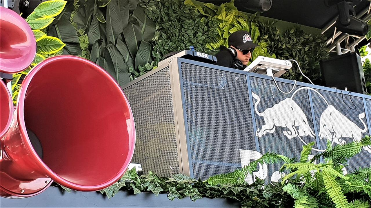 DJ w czapce i słuchawkach miksuje muzykę na stanowisku z logo dwóch byków, otoczonym zielenią i czerwonymi tubami głośników.