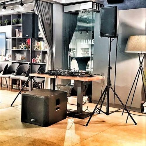 Sprzęt DJski: mikser na drewnianym stole, subwoofer i dwa głośniki na statywach w pomieszczeniu z regałem na książki i lampą podłogową.