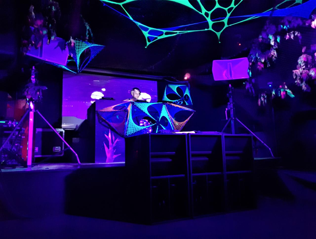 DJ na scenie z geometrycznymi dekoracjami UV, oświetleniem i nagłośnieniem podczas imprezy w klubie.