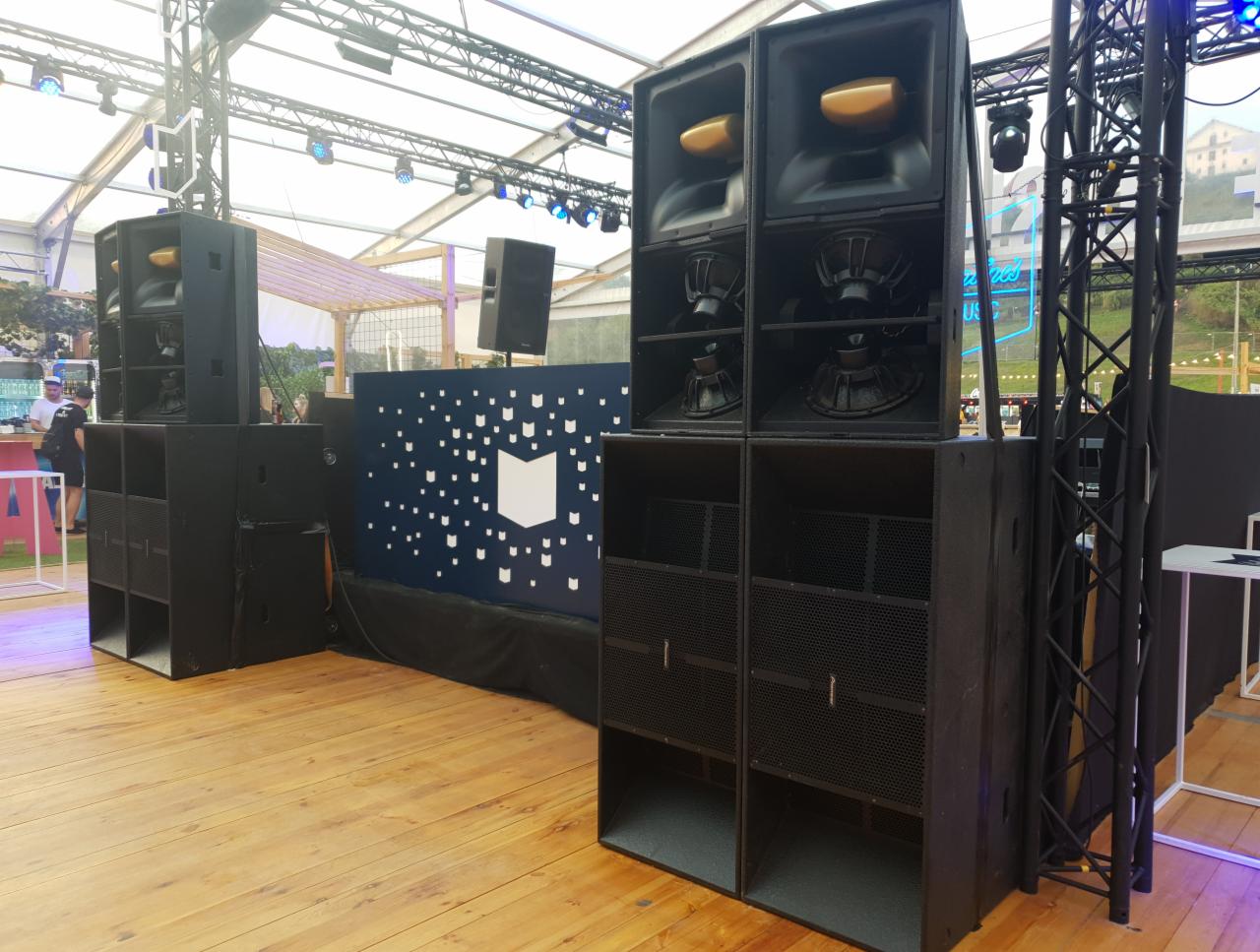 Profesjonalny system nagłośnieniowy na drewnianej scenie, składający się z kolumn głośnikowych i subwooferów, ustawiony przed dekoracyjną ścianką z motywem graficznym.