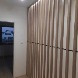 Drewniana ścianka działowa z pionowych listew, podświetlona od góry, z czarnymi, poziomymi łącznikami, widok z korytarza na drewnianą podłogę i drzwi wejściowe.