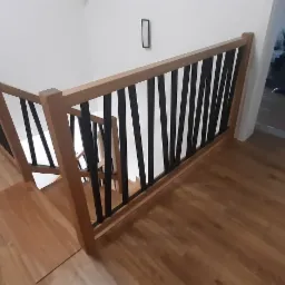 Nowoczesne schody z drewnianą ramą i czarnymi, pionowymi szczeblami balustrady, widoczne z góry na tle drewnianej podłogi i białych ścian.