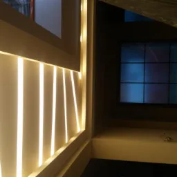 Nowoczesne oświetlenie ścienne z pionowymi pasami LED w jasnym odcieniu, widoczne okno w tle, fragment zabezpieczonej folią podłogi.