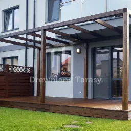 Zadaszony taras drewniany z pergolą z ciemnego drewna, przylegający do nowoczesnego domu z szarą elewacją i balkonem z szklaną balustradą, widok na zadbany trawnik.