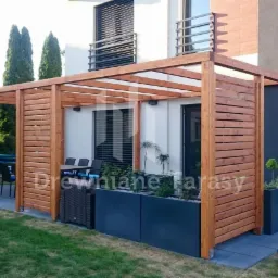 Drewniana konstrukcja pergoli z lamelami, przylegająca do budynku, z meblami ogrodowymi i roślinami w donicach, hamak w tle.