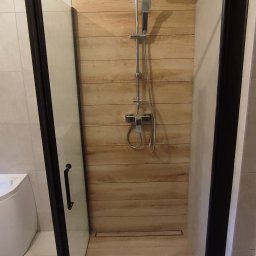 Krzysztof Konig - Kabina prysznicowa z czarnymi ramami, drewnopodobne płytki na ścianie i podłodze, odpływ liniowy, bateria prysznicowa z deszczownicą i słuchawką.