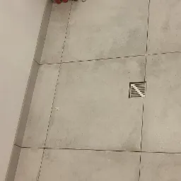 Szare płytki podłogowe w łazience z odpływem liniowym, widoczne rury doprowadzające wodę oraz fragment instalacji kanalizacyjnej w trakcie montażu.