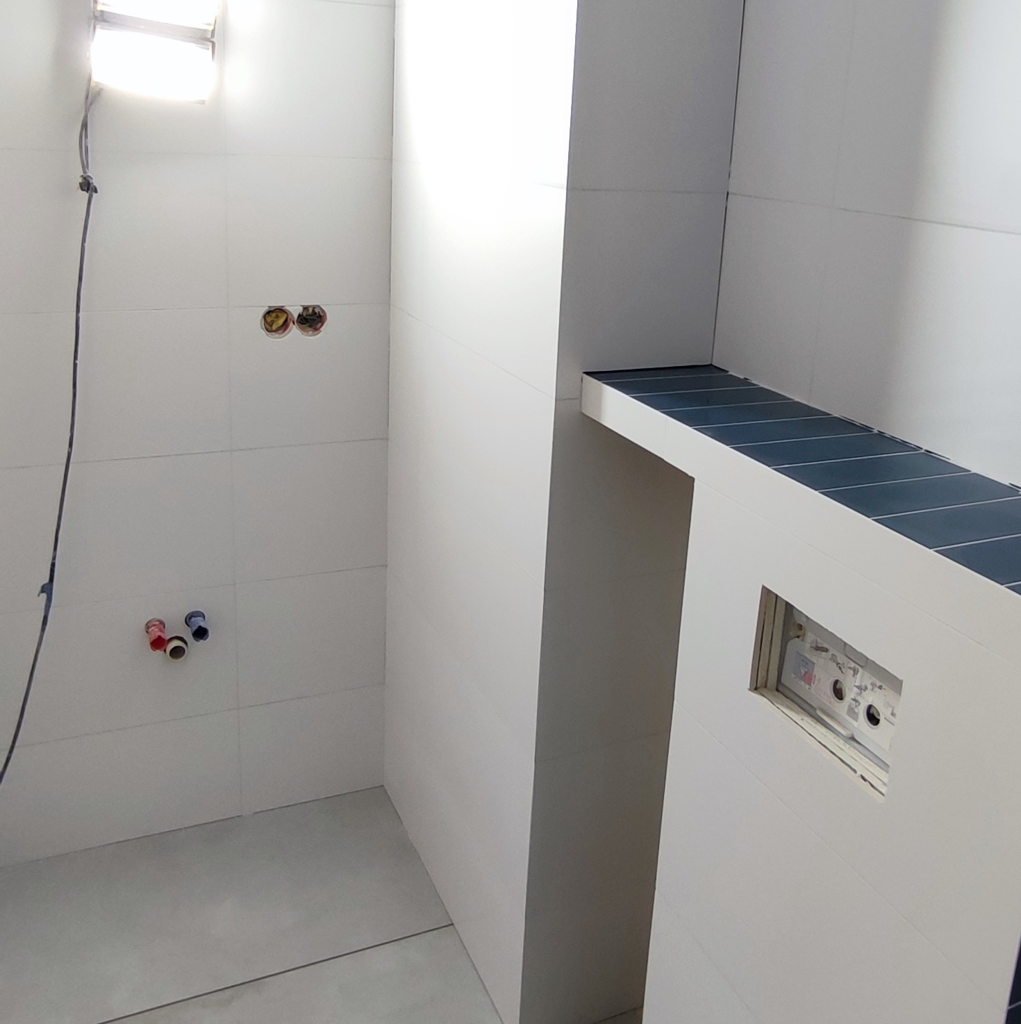Wnętrze łazienki w trakcie prac wykończeniowych: białe płytki ścienne, przygotowane instalacje wodne i elektryczne, podświetlona półka z ciemnymi płytkami oraz otwór na panel sterowania.