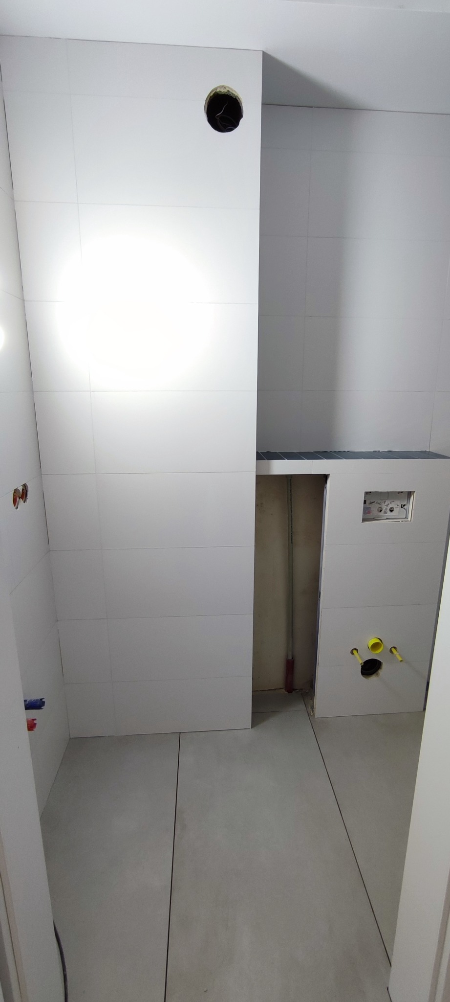 Wnętrze łazienki w trakcie prac wykończeniowych: białe płytki ścienne i podłogowe, widoczne instalacje wodne i elektryczne, otwór w ścianie pod oświetlenie.