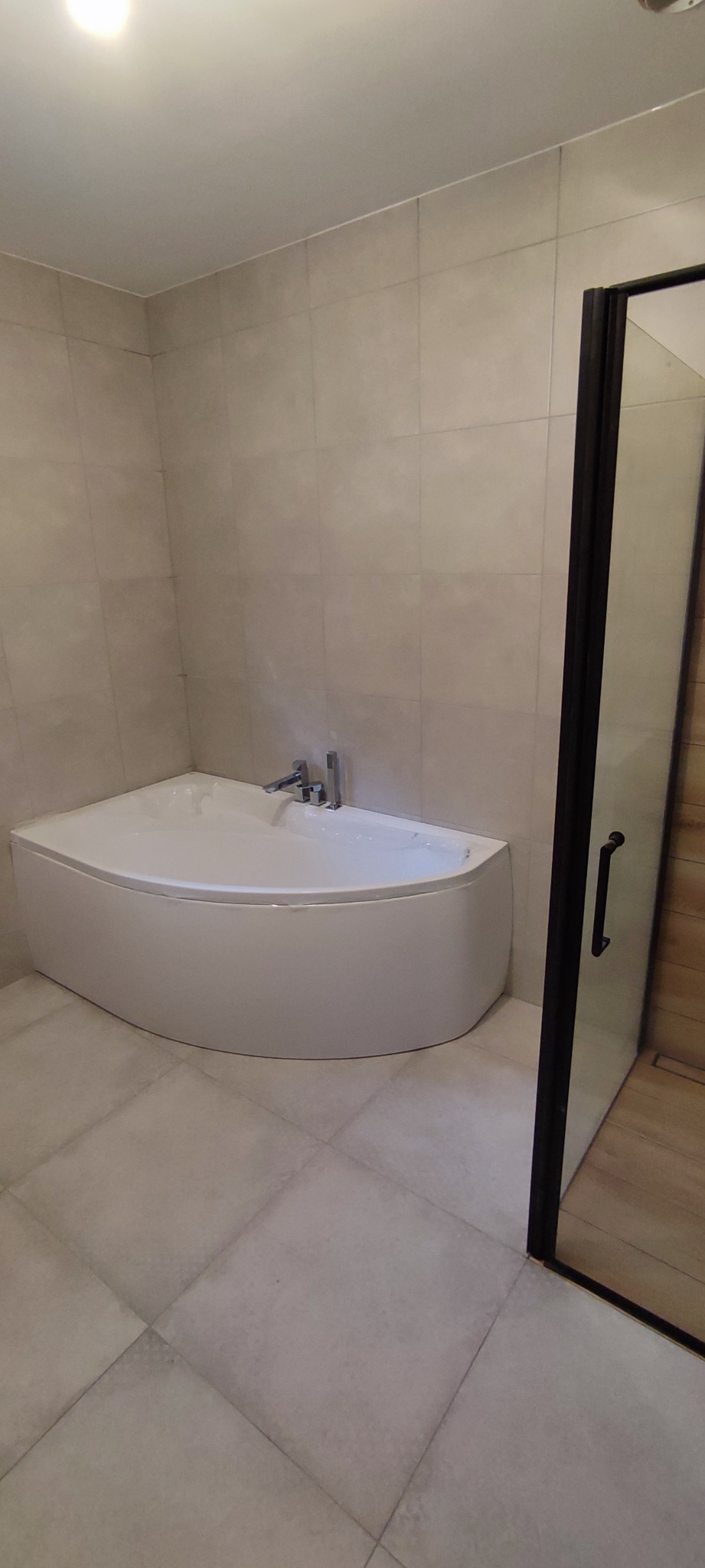 Nowoczesna, narożna wanna akrylowa z chromowaną armaturą, przylegająca do kabiny prysznicowej z czarną ramą, całość wykończona jasnymi płytkami w neutralnej tonacji.