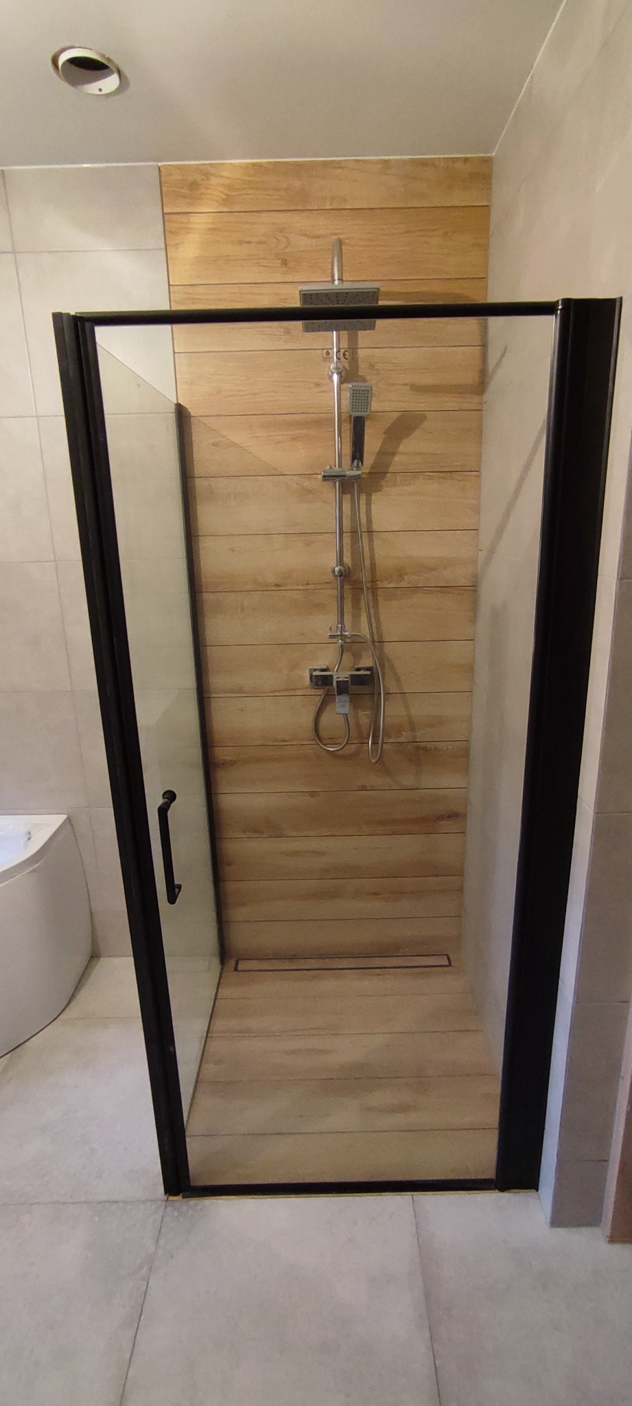Kabina prysznicowa z czarnymi ramami, drewnopodobne płytki na ścianie i podłodze, odpływ liniowy, bateria prysznicowa z deszczownicą i słuchawką.