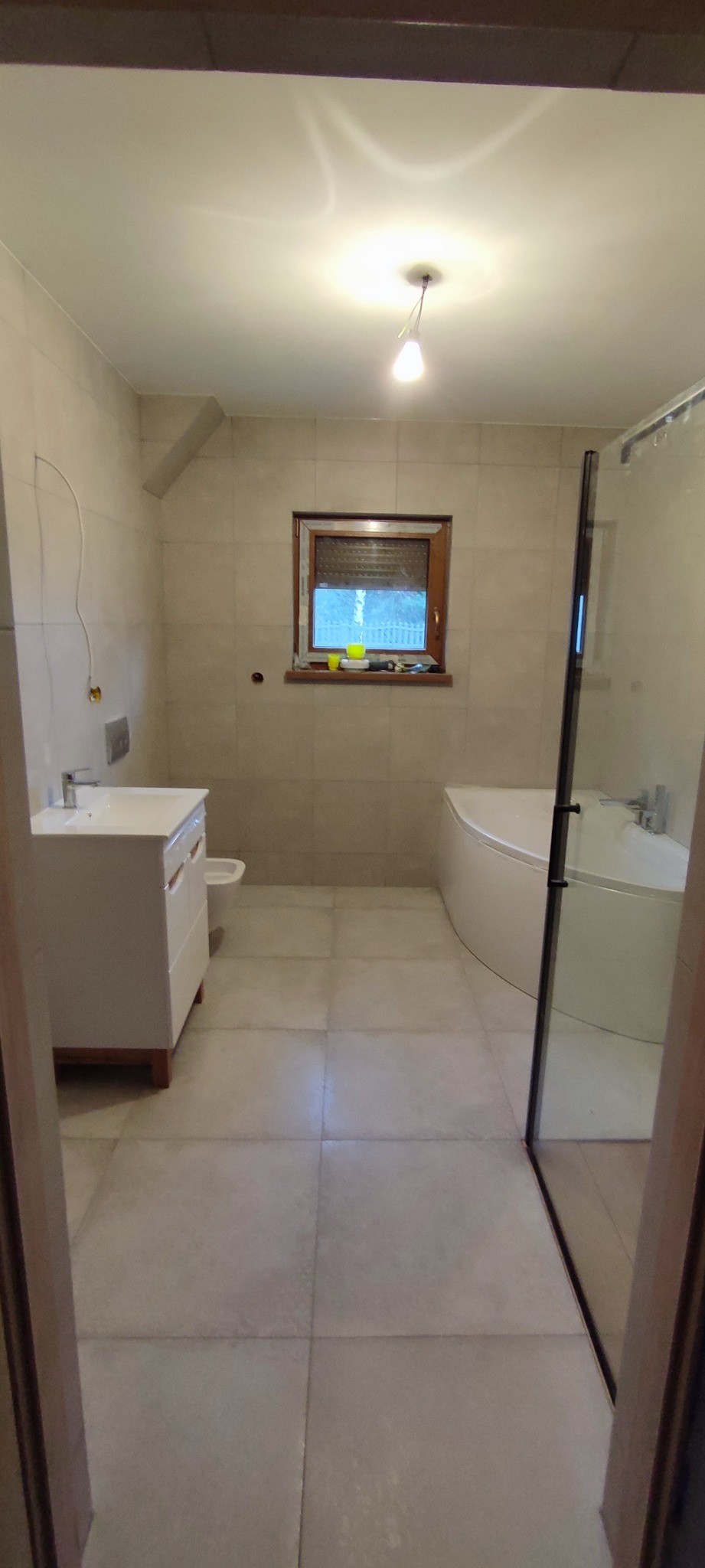 Jasna łazienka z beżowymi płytkami, białą umywalką z szafką, bidetem, narożną wanną i kabiną prysznicową, widok przez framugę drzwi.