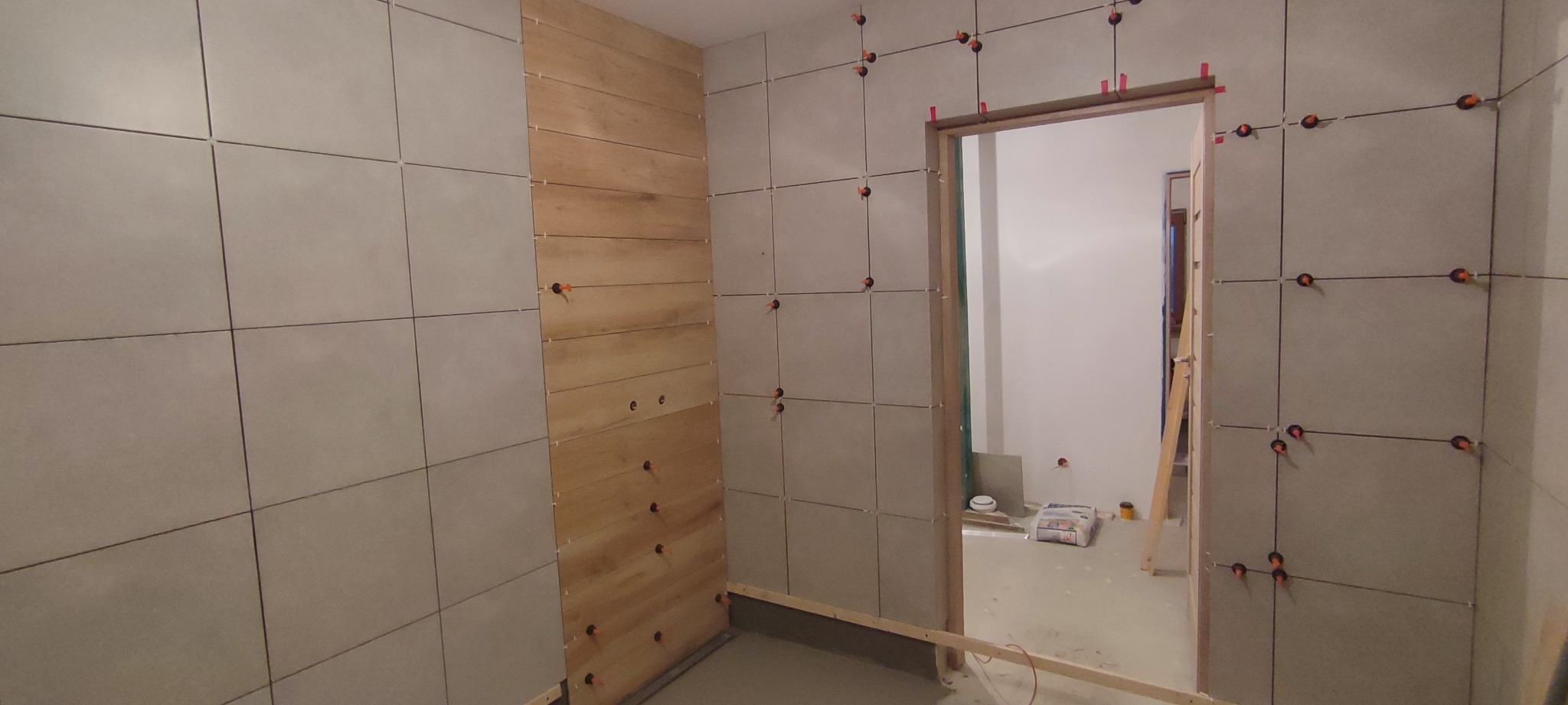 Montaż płytek ceramicznych i drewnopodobnych na ścianie w łazience, widoczne systemy poziomowania płytek i otwory na instalacje.