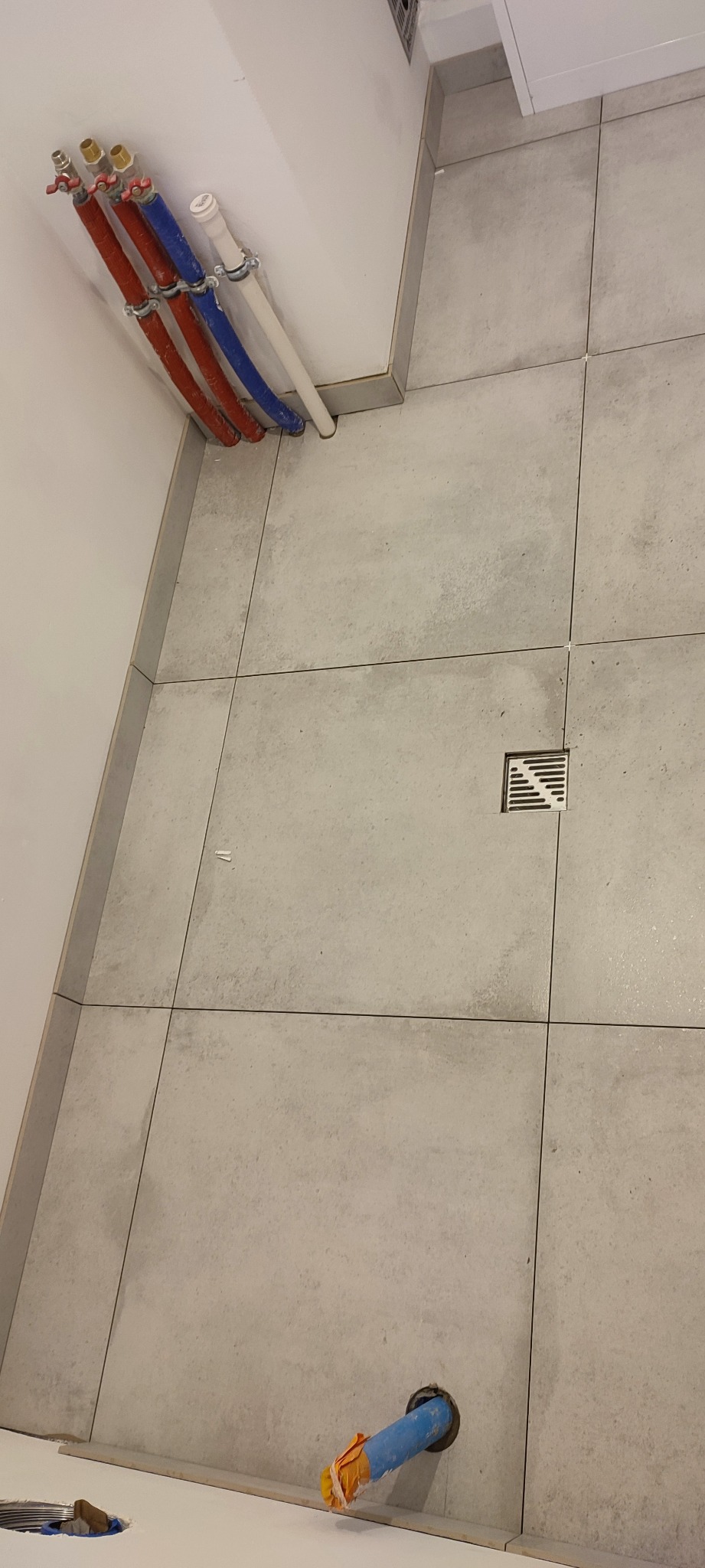 Szare płytki podłogowe w łazience z odpływem liniowym, widoczne rury doprowadzające wodę oraz fragment instalacji kanalizacyjnej w trakcie montażu.