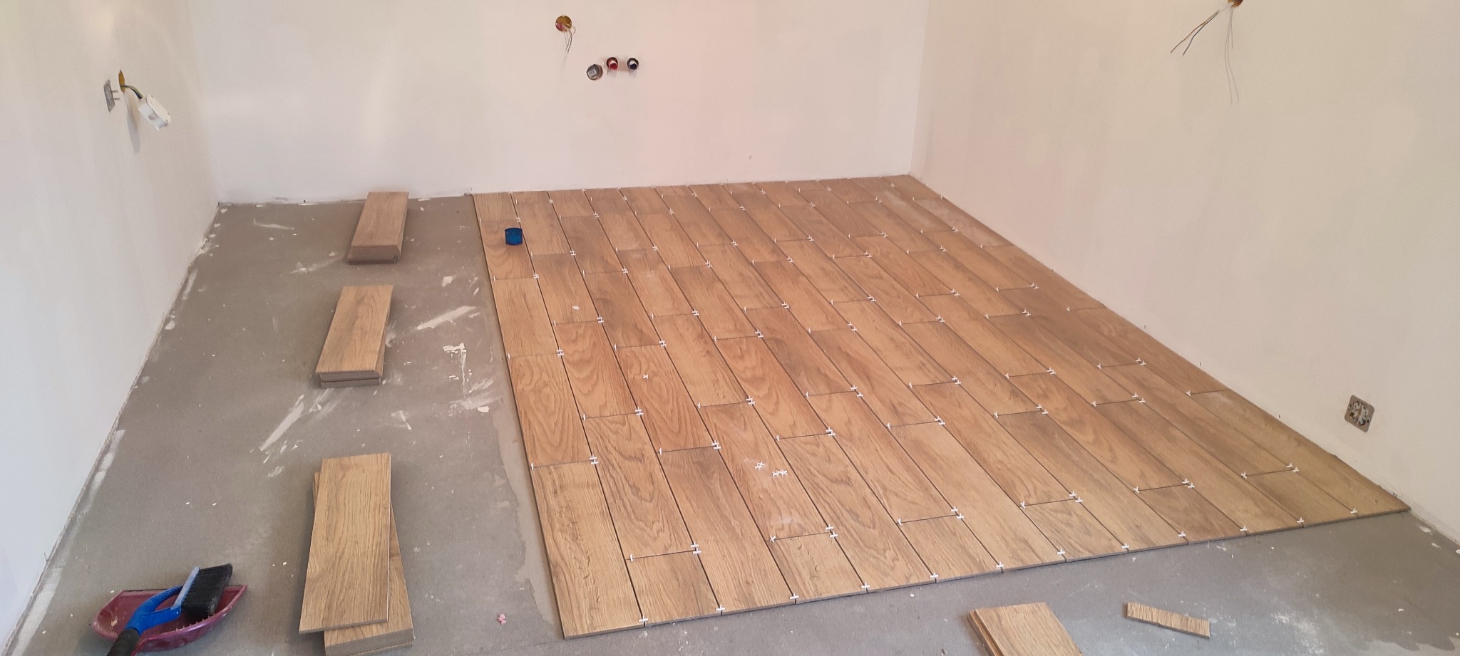 Układanie drewnopodobnych płytek na podłodze w pomieszczeniu, widoczne krzyżyki dystansowe, przygotowane płytki do ułożenia, surowe ściany i instalacja elektryczna.
