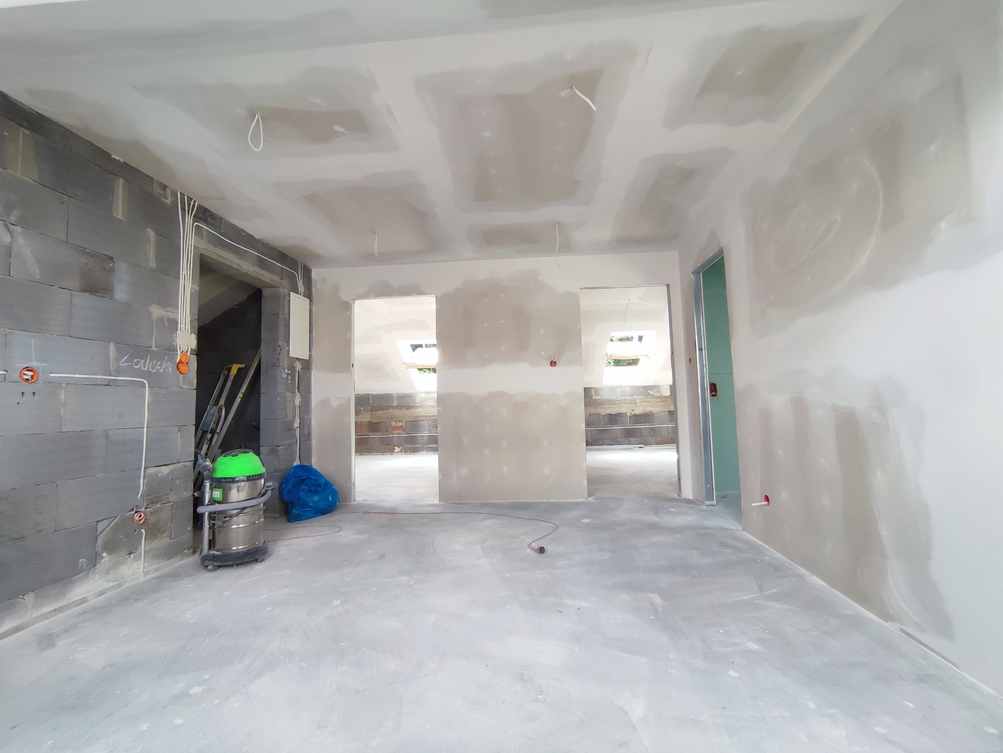 Wnętrze w trakcie prac wykończeniowych: ściany z surowych bloków i częściowo otynkowane, widoczne przewody elektryczne, drabina, odkurzacz przemysłowy i niebieski worek na gruz.