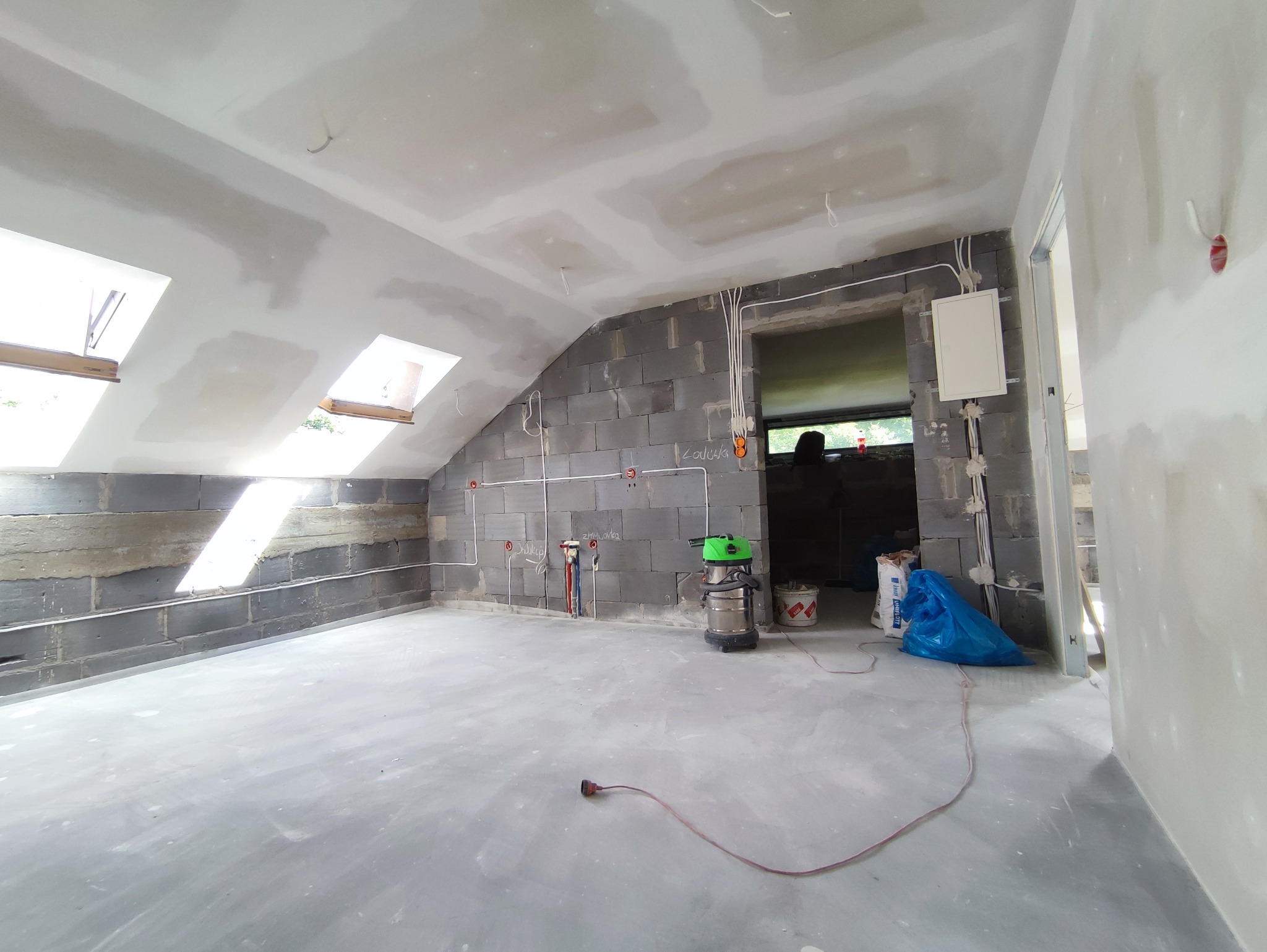 Surowy stan poddasza w trakcie prac wykończeniowych: ściany z szarego betonu komórkowego, częściowo otynkowane, z widocznymi instalacjami elektrycznymi i hydraulicznymi, dwa okna dachowe w skośnym...