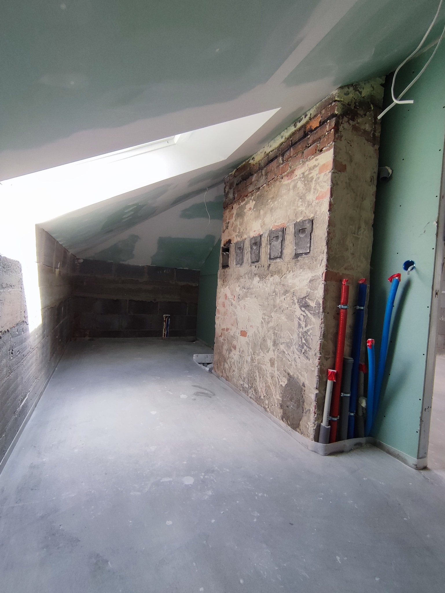 Surowa łazienka na poddaszu w trakcie remontu: widoczne instalacje wodne, fragment ceglanej ściany z metalowymi klapami rewizyjnymi, sufit i ściany pokryte płytami gipsowo-kartonowymi, okno dachowe.
