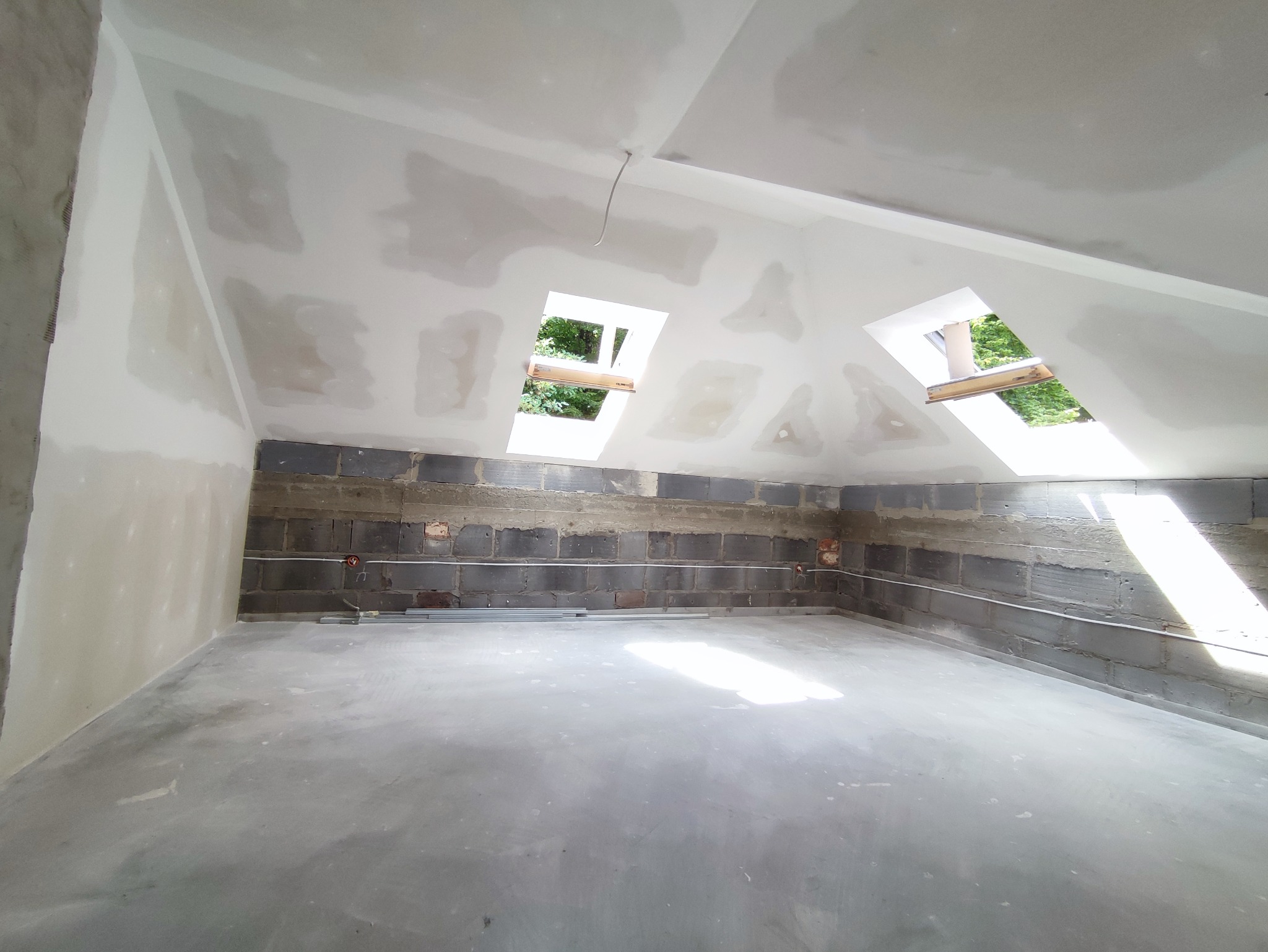 Poddasze w trakcie adaptacji: ściany i sufit pokryte gładzią szpachlową, widoczne dwa okna dachowe z widokiem na zieleń, ściana z bloczków częściowo przygotowana pod wykończenie, wystające przewody...