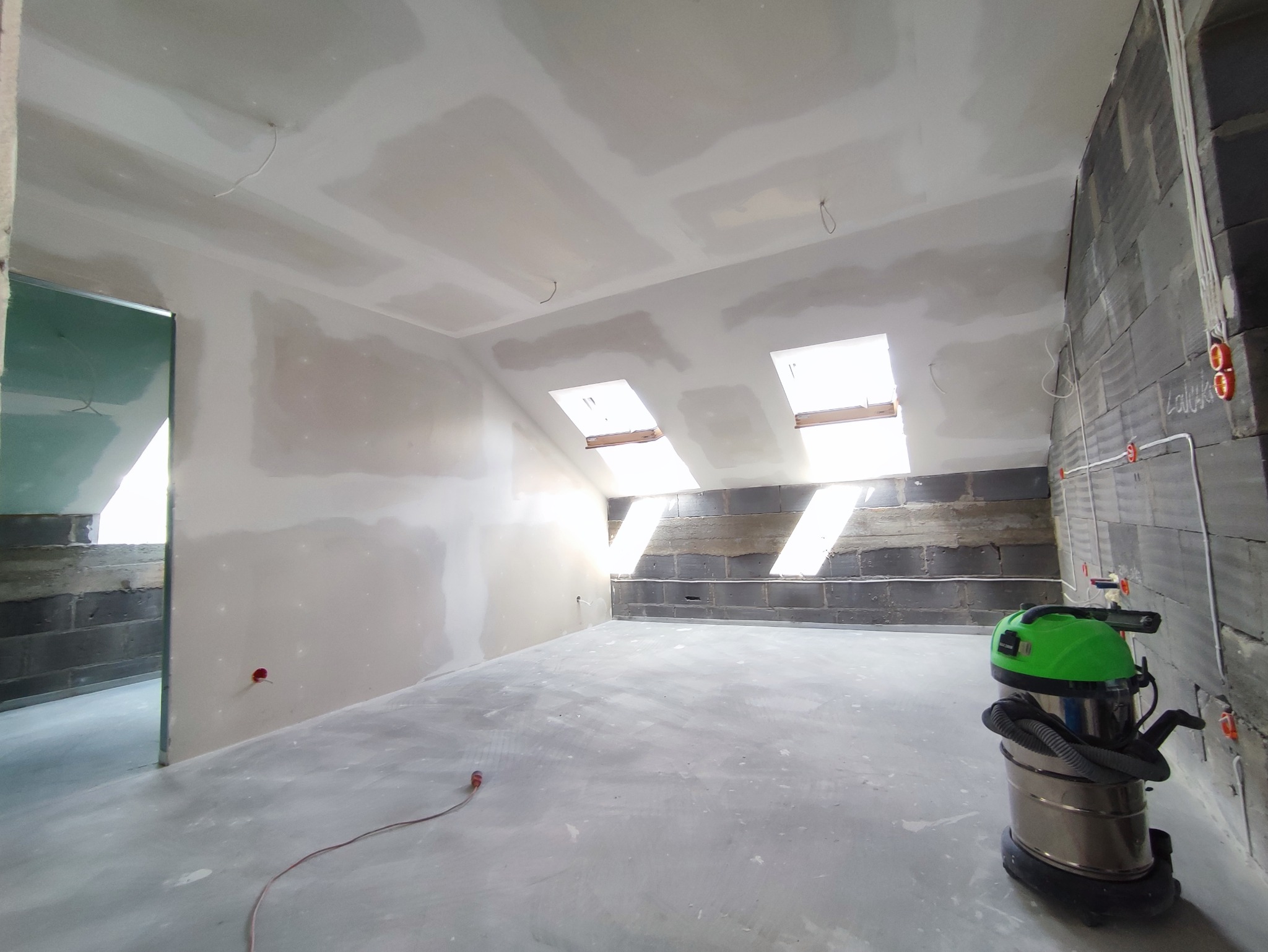 Poddasze w trakcie wykańczania: ściany z płyt gipsowo-kartonowych częściowo wyszpachlowane, surowa ściana z bloczków betonowych z instalacją elektryczną, widoczne okna dachowe i odkurzacz przemysłowy.