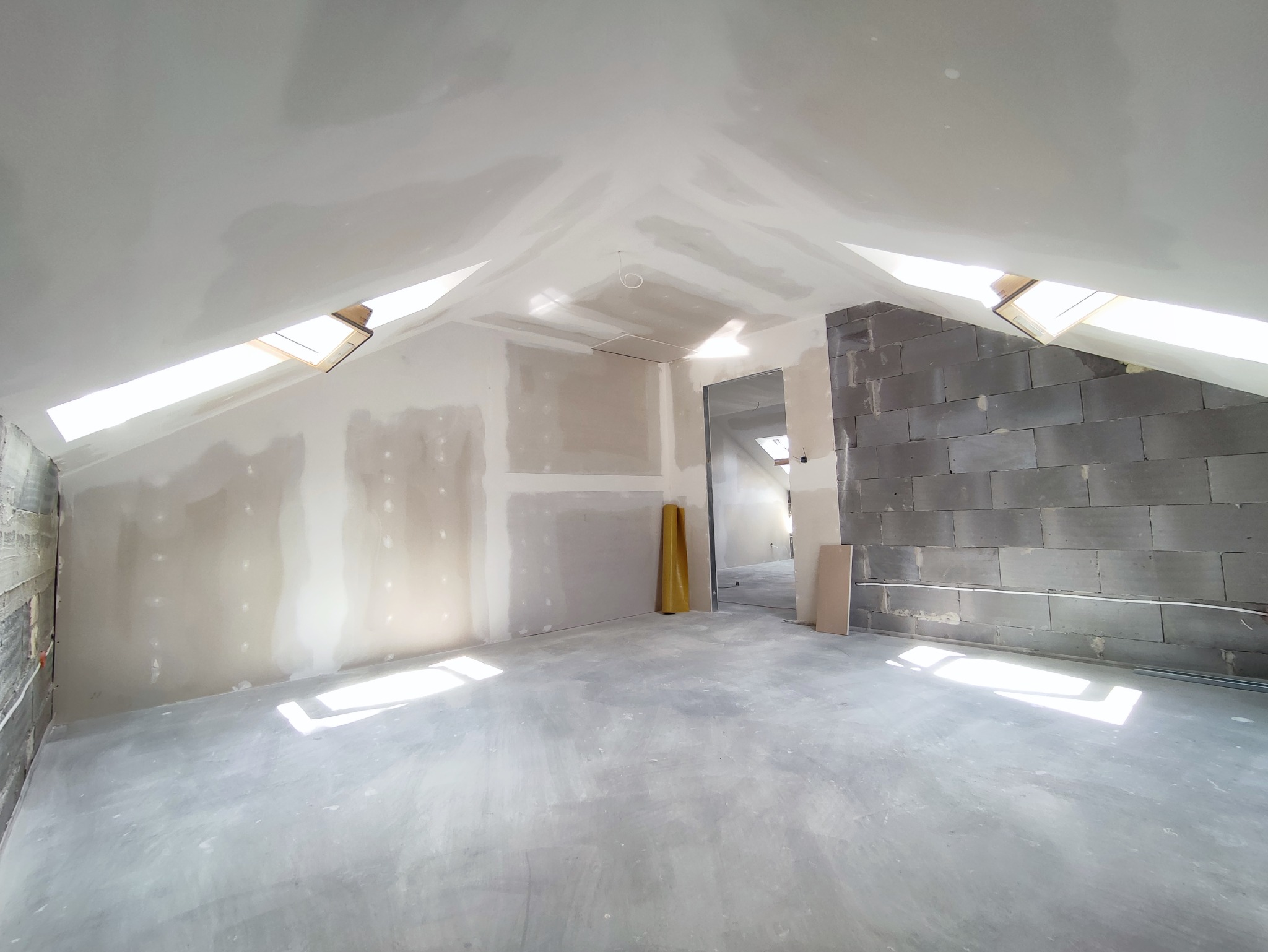 Poddasze w trakcie remontu: ściany z płyt gipsowo-kartonowych częściowo zaszpachlowane, fragment ściany z szarego bloczka, okna dachowe w skosach, żółta folia budowlana przy ścianie.