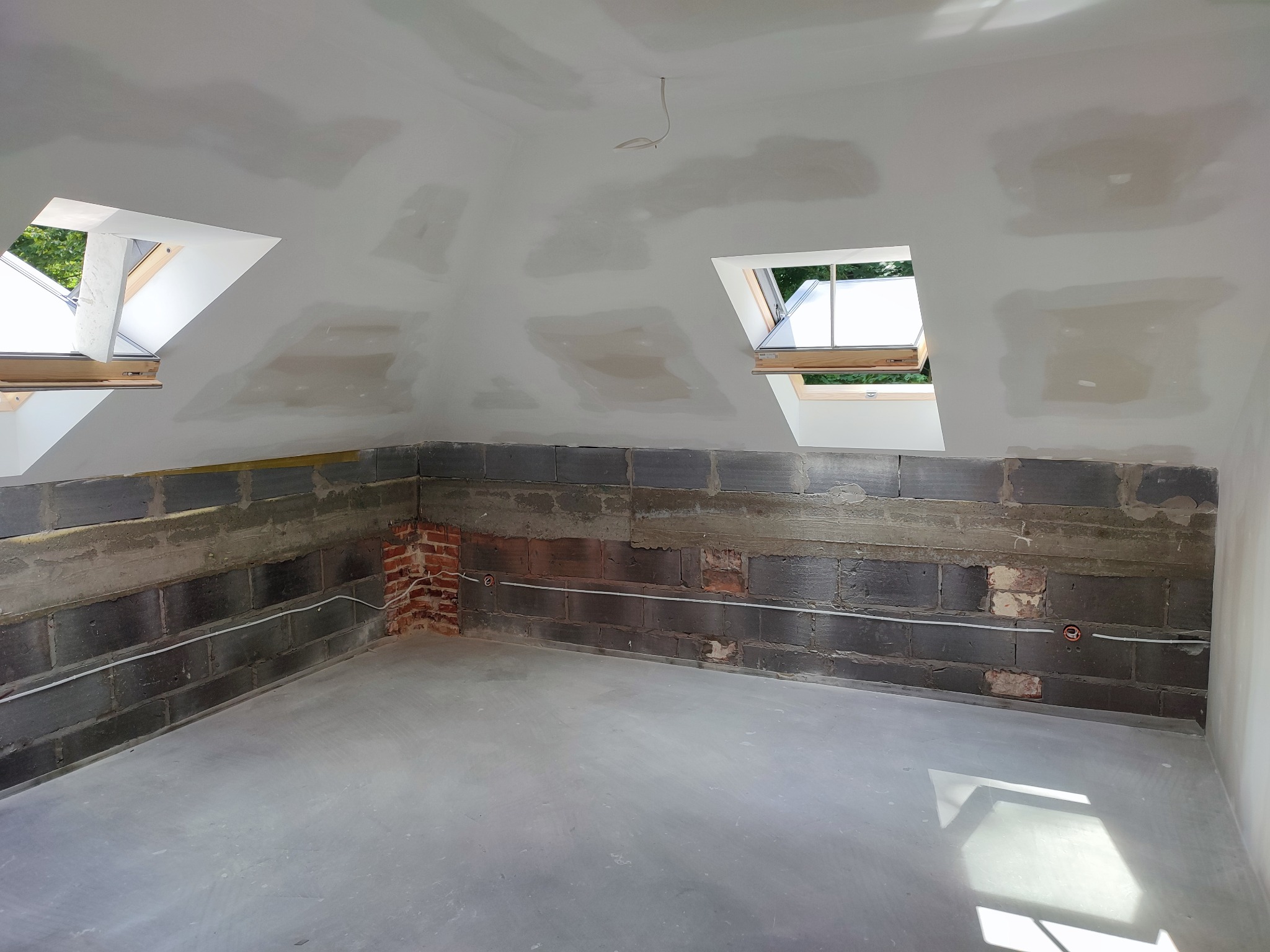 Surowe wnętrze poddasza w trakcie prac wykończeniowych: ściany z bloków betonowych, częściowo otynkowane, z widoczną instalacją elektryczną, gładki betonowy wylewka na podłodze, dwa okna dachowe...