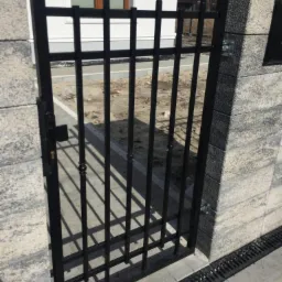 Czarna, metalowa furtka z ozdobnymi grotami, zamontowana między dwoma słupkami z szarego betonu architektonicznego, widok z bliska na tle fragmentu domu.