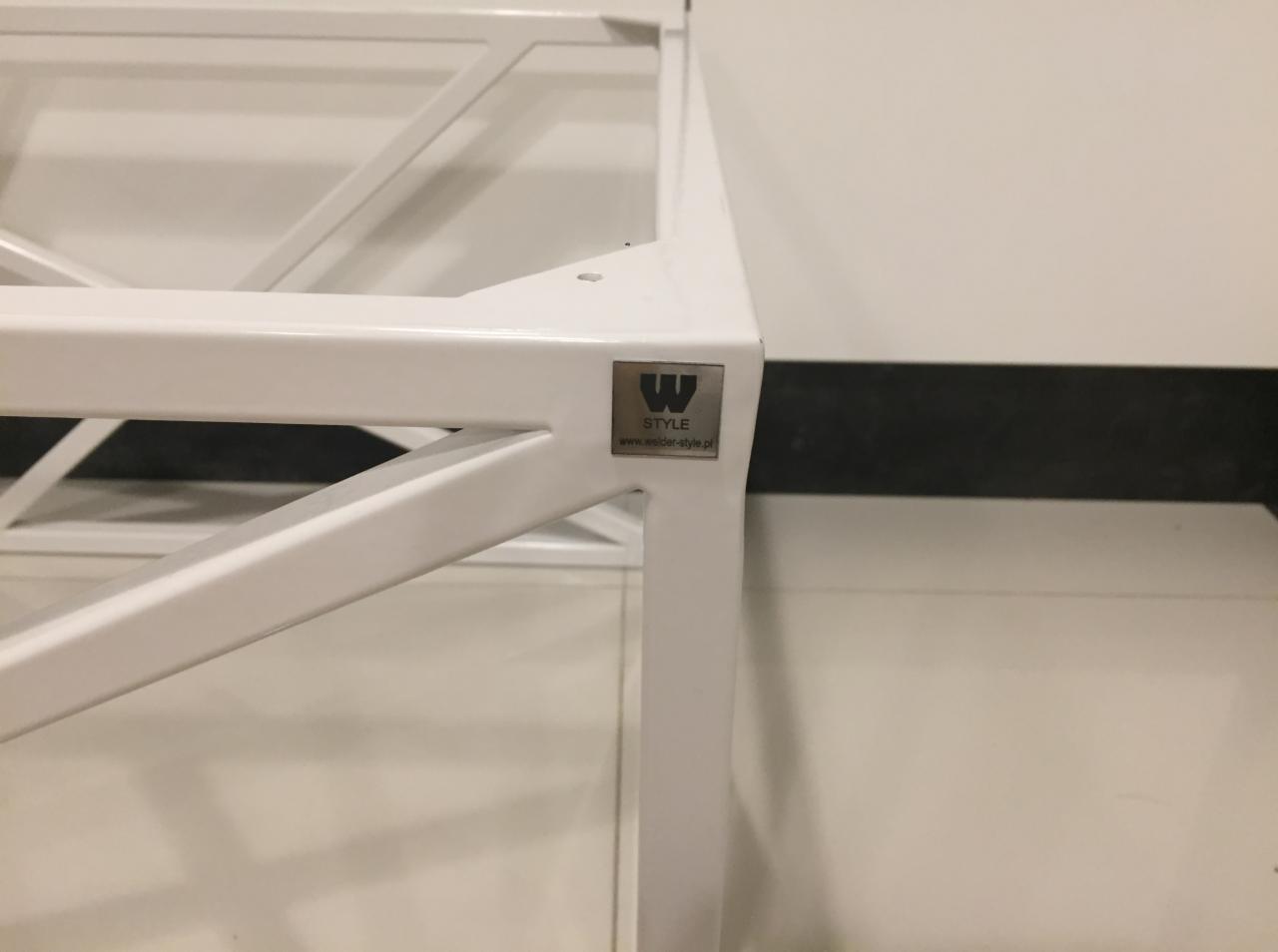 Fragment białej, metalowej konstrukcji schodów z naklejoną srebrną etykietą z logo i adresem strony internetowej producenta.