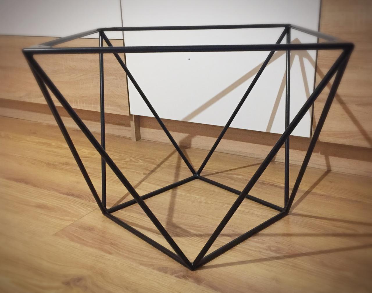 Geometryczna konstrukcja z czarnych, metalowych prętów tworząca ażurową bryłę na drewnianej podłodze, częściowo widoczna biała szafka w tle.