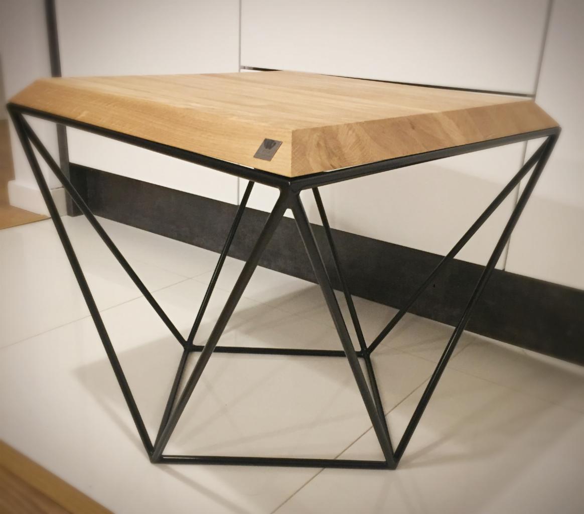 Stolik kawowy z blatem z jasnego drewna i geometryczną podstawą z czarnych, metalowych prętów, na białej podłodze.