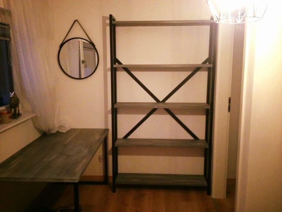 Regał z metalową ramą i półkami z szarego drewna, stojący obok biurka z blatem w tym samym stylu, okrągłe lustro na ścianie, w tle okno z zasłoną.