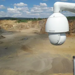 Biała kamera przemysłowa Hikvision zamontowana na zewnątrz z widokiem na rozległy, piaszczysty teren kamieniołomu pod błękitnym niebem z chmurami.