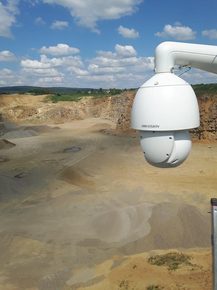 Biała kamera przemysłowa Hikvision zamontowana na zewnątrz z widokiem na rozległy, piaszczysty teren kamieniołomu pod błękitnym niebem z chmurami.