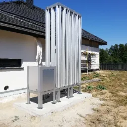 Zewnętrzna jednostka dużej pompy ciepła powietrze-woda z pionowymi rurami aluminiowymi, zamontowana na betonowej podstawie obok domu jednorodzinnego z czarnym dachem, paleta drewniana na pierwszym...