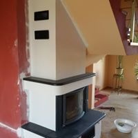 Biało-czarny kominek z prostokątną obudową, widoczne niedokończone wykończenie ściany w kolorze pomarańczowym i częściowo pomalowane schody, mała miska i narzędzia leżące na podłodze.