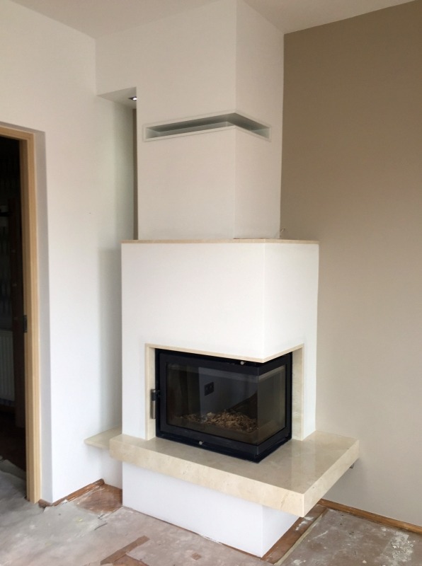 Nowoczesny kominek z czarnym wkładem, obudowany białym tynkiem i marmurowym blatem, w trakcie montażu, widoczne niedokończone wykończenie podłogi.