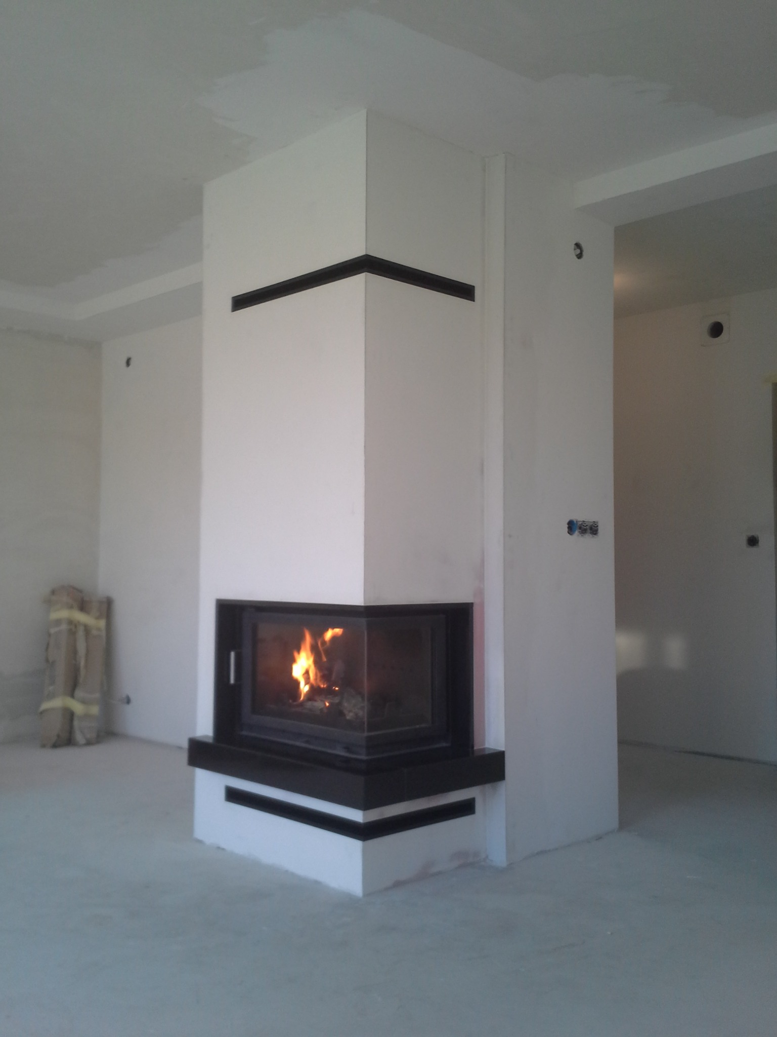 Nowoczesny narożny kominek z czarnymi akcentami w surowym, wykończonym na biało wnętrzu, z widocznym płomieniem i workami z zaprawą w tle.