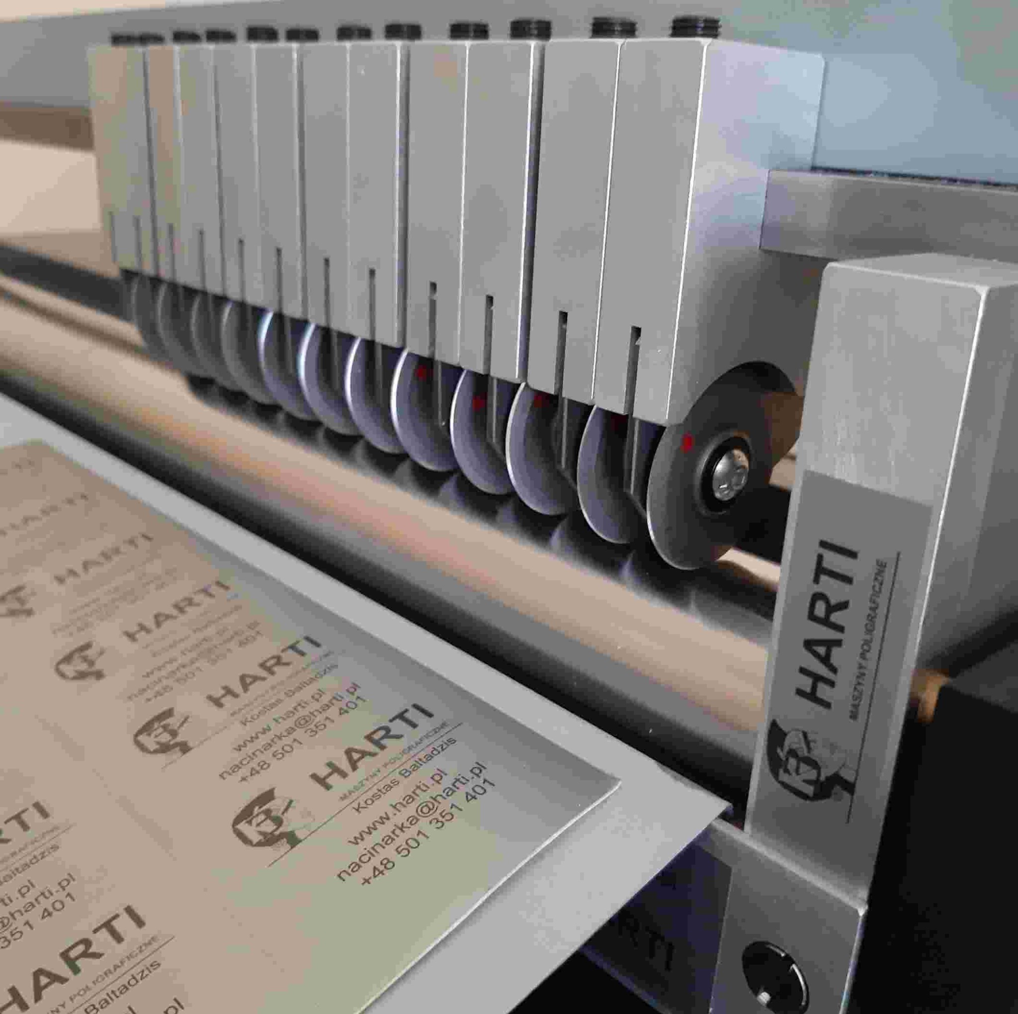 Maszyna poligraficzna tnąca arkusz z wizytówkami firmy HARTI. Widoczne noże tnące i logo firmy na wizytówkach. Precyzyjna obróbka materiałów.