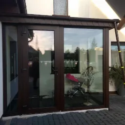 Zabudowa tarasu z brązowymi ramami, widoczne drzwi balkonowe i okno, wewnątrz wózek dziecięcy i dekoracje, na zewnątrz kostka brukowa.