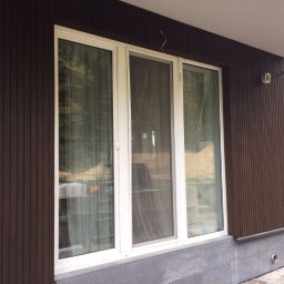 Instroybel Sp. z o.o. - Elewacja domu z brązowymi panelami pionowymi, trzema białymi oknami i szarą podmurówką, widoczne okablowanie elektryczne na zewnątrz.