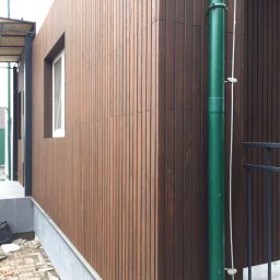 Instroybel Sp. z o.o. - Elewacja budynku obłożona pionowymi panelami z drewna o ciepłym brązowym odcieniu, z widocznym oknem i zieloną rurą spustową, ujęcie z niskiego kąta.