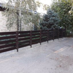 Instroybel Sp. z o.o. - Nowoczesne, poziome ogrodzenie z ciemnego drewna, zamontowane na betonowej podmurówce, widoczne z perspektywy chodnika, z budynkiem w tle i drzewami po obu stronach.