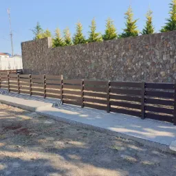 Nowoczesne, poziome, brązowe ogrodzenie panelowe z betonu architektonicznego, z kamiennym murem w tle i młodymi iglakami, w słoneczny dzień.