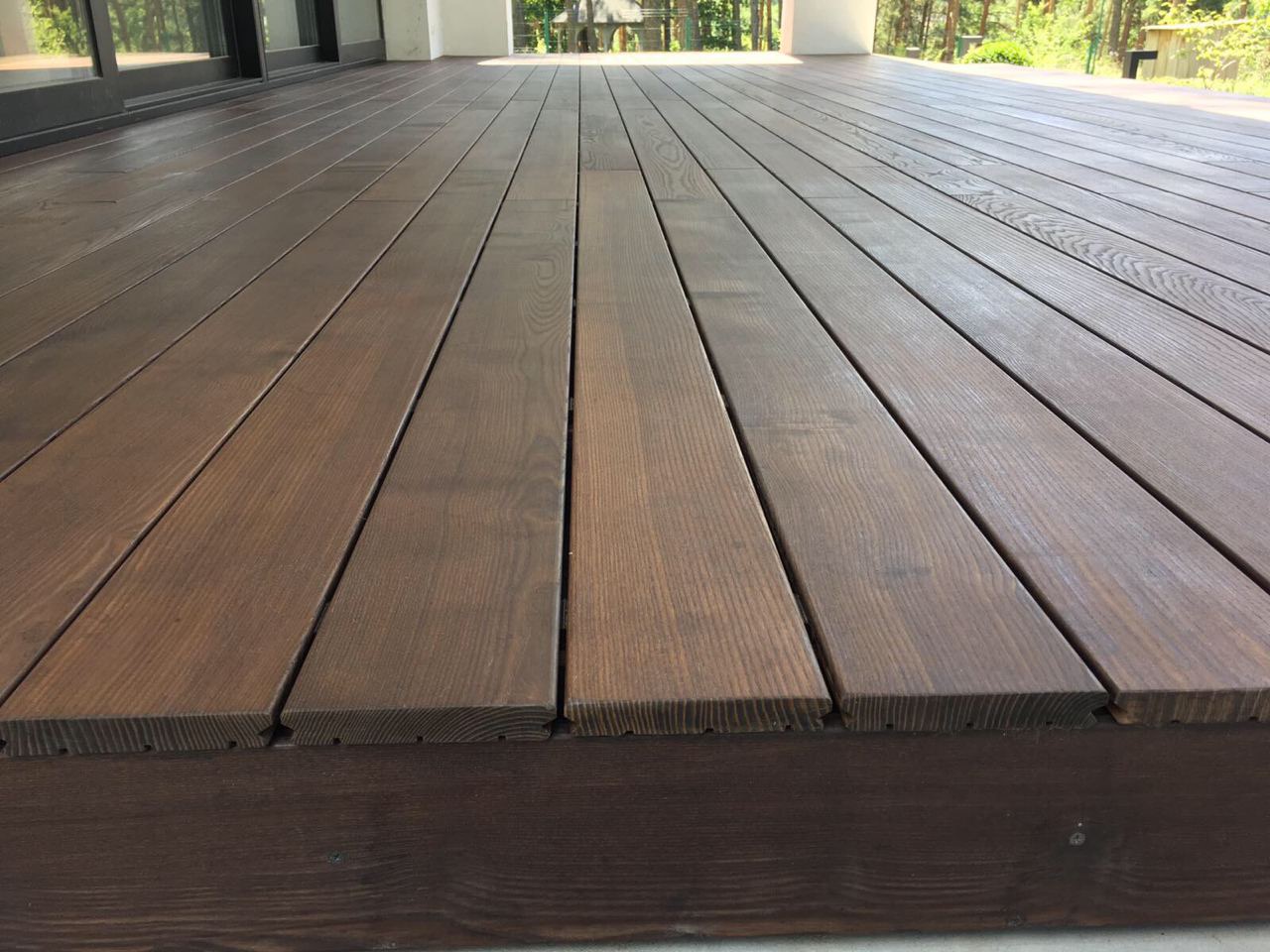 Nowoczesny taras z ciemnobrązowych desek, z widocznymi słojami drewna, prowadzący do przeszklonych drzwi balkonowych i białych kolumn, otoczony zielenią drzew.