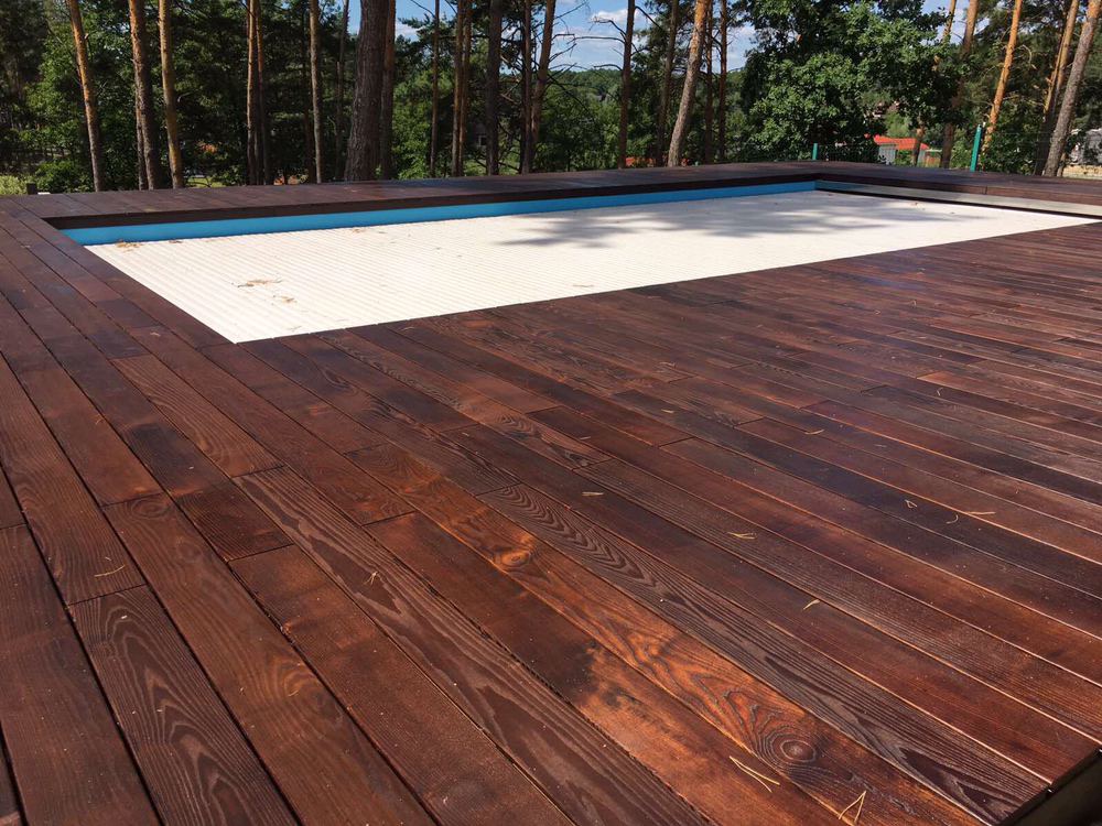 Ciemnobrązowy taras z desek otaczający basen z białą roletą, w tle drzewa iglaste.