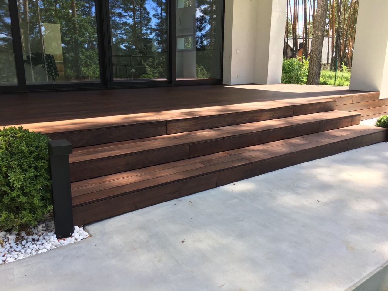 Nowoczesny taras z ciemnego drewna z trzema stopniami, zintegrowany z budynkiem, oświetlony lampą ogrodową i otoczony białymi kamykami.