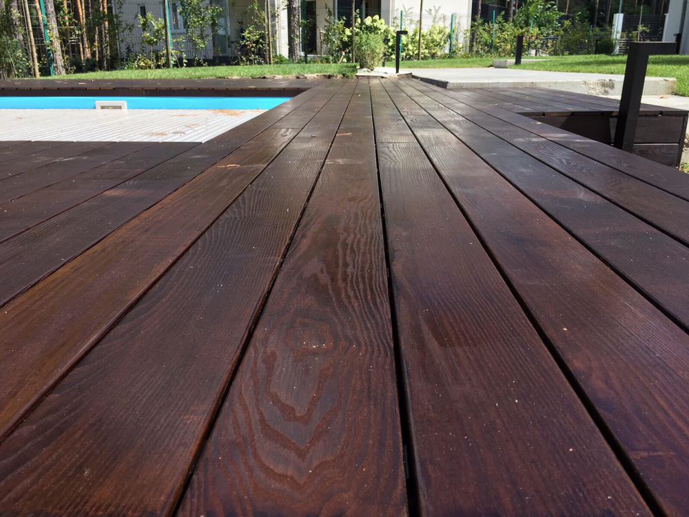 Ciemnobrązowy taras z desek ułożonych równolegle, przylegający do basenu z częściowo zasłoniętą powierzchnią, widok z niskiej perspektywy.