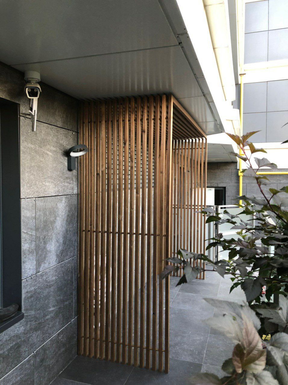 Nowoczesna, drewniana przesłona z pionowych listew, zamontowana na tarasie z szarych płyt, z widoczną kamerą monitoringu na ścianie budynku w tle.