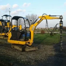 Żółta mini koparka JCB z wiertnicą hydrauliczną na nierównym terenie, w tle druga koparka i biały bus dostawczy, drzewa i błękitne niebo.